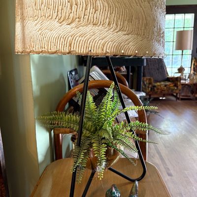 Mid-century modern tripod table lamp featuring a black metal frame, integrated brass planter with an artificial fern, and a textured...