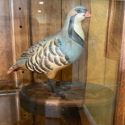 Hand-carved and painted wooden partridge decoy figurine mounted on a circular wooden base, displayed inside a glass cabinet.