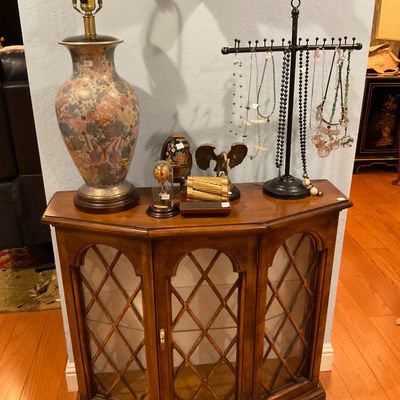 Vintage wooden console cabinet with glass doors and diamond-patterned wood overlays, displaying a floral lamp. 