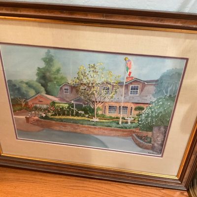 Framed watercolor painting depicting a suburban house with a flagpole, signed by the artist, housed in a wood frame with a fabric mat.