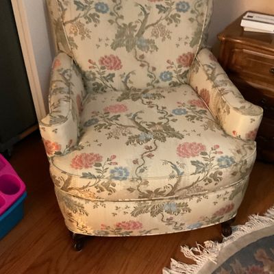 Vintage upholstered armchair featuring a floral and vine pattern in pink, blue, and green on a cream background, supported by dark wood...