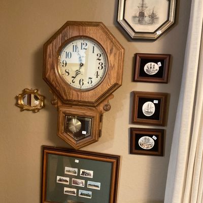Vintage oak regulator-style wall clock with a pendulum, displayed alongside framed nautical artwork and a collection of train illustrations.