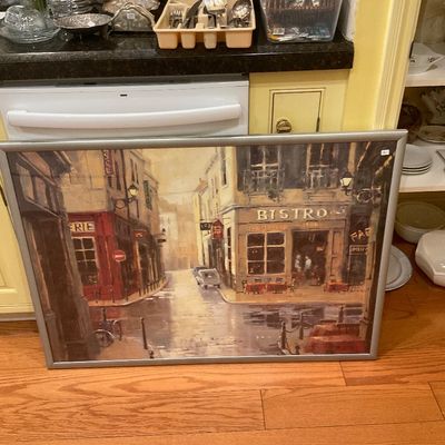 Large framed print depicting a European street scene with a bistro, set in a simple silver-toned metal frame.