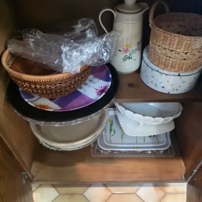 Sale Photo Thumbnail #52: Assorted kitchenware and serving items inside a wooden cabinet, including woven baskets, a floral pitcher, decorative plates, and milk glass bowls.