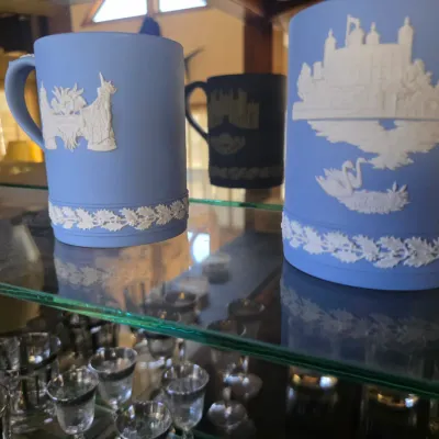 Sale Photo Thumbnail #24: Wedgwood light blue jasperware mugs featuring white relief designs of historic English landmarks, displayed on a glass shelf.