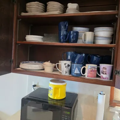 Sale Photo Thumbnail #63: Assorted kitchenware including ceramic mugs, plates, and bowls organized in a wooden cabinet above a black countertop microwave.