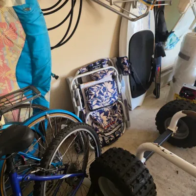 Sale Photo Thumbnail #99: Assorted outdoor and beach gear, including a blue Trek bicycle, a folded floral beach chair, a surfboard, and a small utility trailer with knobby tires.