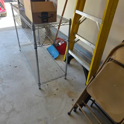 Sale Photo Thumbnail #104: Three-tier chrome wire shelving unit, a yellow fiberglass stepladder, and a stack of beige metal folding chairs in a garage setting.
