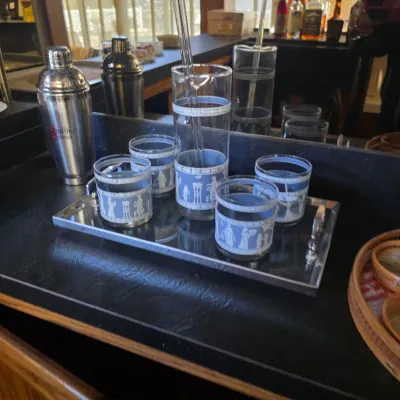 Sale Photo Thumbnail #25: Vintage mid-century barware set featuring a blue and white Greek key and classical figure motif, including four lowball glasses and a matching pitcher on a mirrored tray.