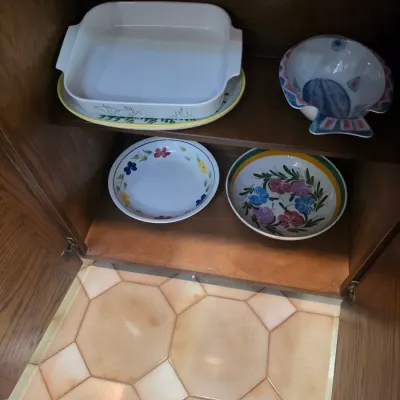 Sale Photo Thumbnail #53: Assorted ceramic serving dishes, including a white rectangular baking dish, a fish-shaped bowl, and two floral-patterned bowls, displayed inside a wooden cabinet.