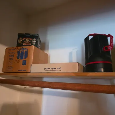 Sale Photo Thumbnail #6: Assorted closet shelf items including a vintage Tri-State revolving poker rack in its original box, a Jack Daniel's tin, a box labeled "Camera System Info," and two portable lanterns.