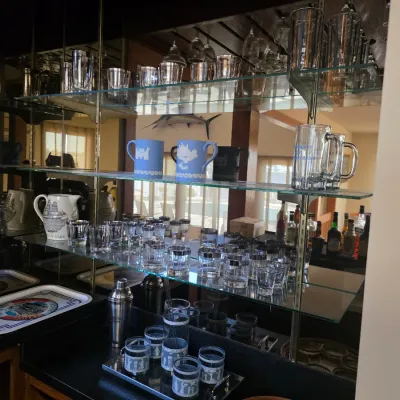 Sale Photo Thumbnail #23: Assorted vintage glassware and barware, including Wedgwood Jasperware mugs, silver-rimmed lowball glasses, and a cocktail shaker, displayed on glass shelves.