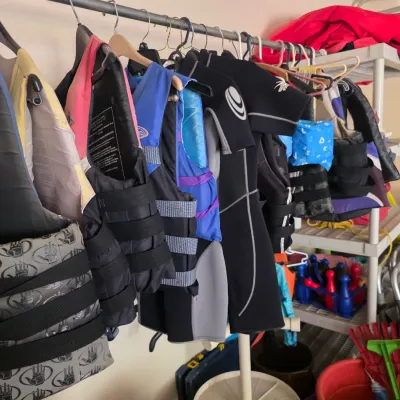 Sale Photo Thumbnail #87: Assorted water sports gear, including life jackets and wetsuits, hanging on a metal rack.