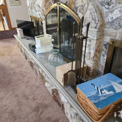 Sale Photo Thumbnail #33: Assorted home decor and fireplace accessories, including a brass tool set, a lighthouse-themed picnic basket, and a glass jar, arranged on a stone fireplace hearth.