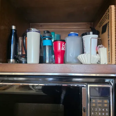 Sale Photo Thumbnail #55: Assorted drinkware, including travel mugs, water bottles, and a plastic pitcher, stored on a shelf above a vintage microwave.