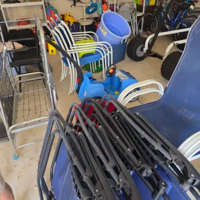 Sale Photo Thumbnail #109: Assorted outdoor and recreational items in a garage, including folding lawn chairs, a blue plastic ride-on toy car, stackable patio chairs, and life jackets.