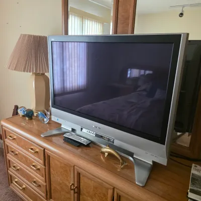Sale Photo Thumbnail #75: Panasonic flat-screen television with silver bezel and stand, resting on a vintage wooden dresser alongside a ceramic table lamp and decorative glass figurines.