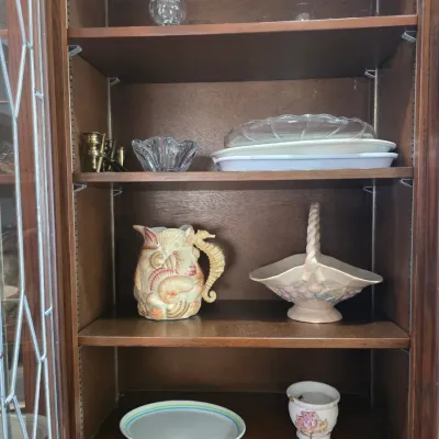 Sale Photo Thumbnail #50: Assorted glassware and ceramic serving pieces, including a seashell-themed pitcher and a ceramic basket, displayed inside a wooden hutch.