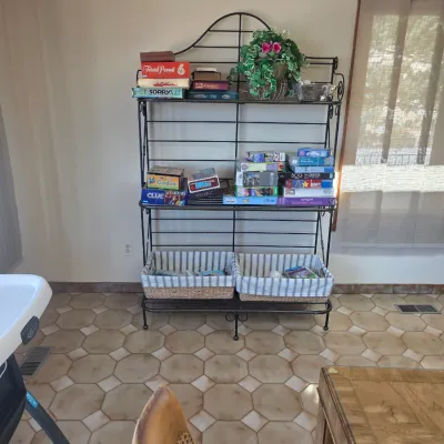Sale Photo Thumbnail #44: Black wrought iron baker's rack with wire shelves, holding an assortment of board games, puzzles, and two fabric-lined wicker storage baskets on the bottom shelf.