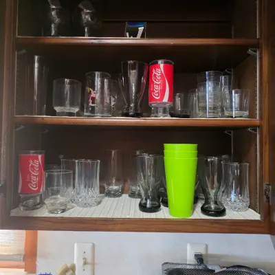 Sale Photo Thumbnail #62: Assorted glassware collection in a wooden cabinet, including Coca-Cola glasses, clear tumblers, and bright green plastic cups.