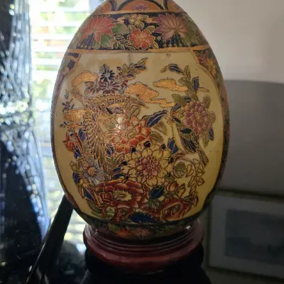 Sale Photo Thumbnail #6: Satsuma-style decorative porcelain egg featuring intricate floral and bird motifs with gold gilt accents, displayed on a dark wooden stand.