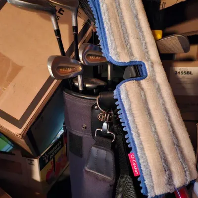 Sale Photo Thumbnail #24: Vintage golf bag containing a set of golf clubs, including Relief irons and woods, shown alongside an O-Cedar microfiber mop in a storage area.