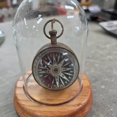 Sale Photo Thumbnail #36: Vintage pocket compass with an ornate compass rose dial, displayed hanging inside a glass dome on a wooden base.