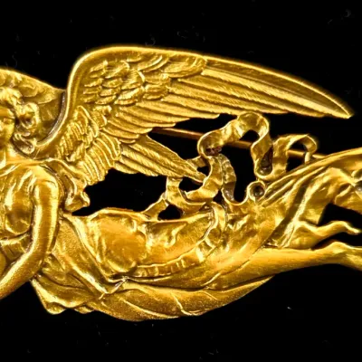 Sale Photo Thumbnail #65: Art Nouveau style gold-tone metal brooch depicting a flying angel with flowing drapery, set against a black background.