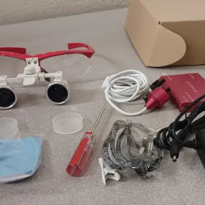 Sale Photo Thumbnail #31: Dental surgical binocular loupes with red frames, accompanied by an LED headlight kit, power adapter, charging cable, screwdriver, and cleaning cloth.