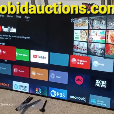 Sale Photo Thumbnail #36: Sony Bravia smart television displaying its home screen, shown with its remote control and stand legs on the floor.