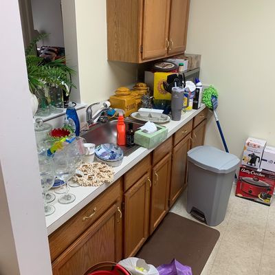Sale Photo Thumbnail #20: Assorted kitchenware and household items, including glassware, ceramic canisters, a Betty Crocker slow cooker in its box, and a grey step-on trash can, arranged on and around oak kitchen cabinets.