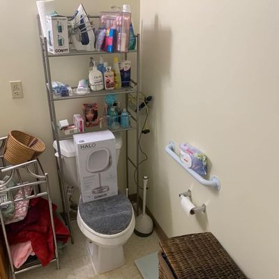 Sale Photo Thumbnail #16: Metal over-the-toilet shelving unit holding various toiletries, a Conair fabric steamer, and an iron, alongside a boxed Conair Halo lighted mirror resting on the toilet seat.