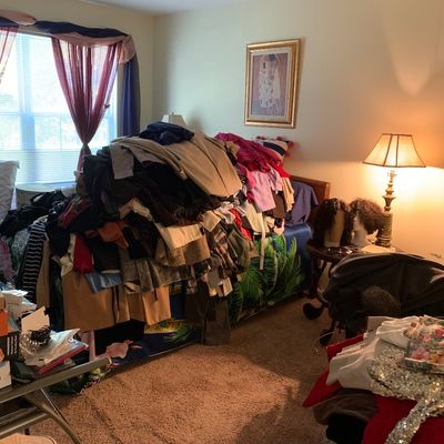 Sale Photo Thumbnail #23: Large assortment of women's clothing piled on a bed, alongside bedroom furniture, a brass table lamp, and framed wall art.