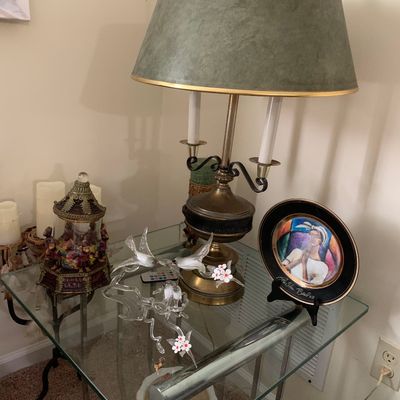 Sale Photo Thumbnail #9: Vintage brass table lamp with a green shade, displayed on a glass-top end table alongside a decorative collector plate, a glass bird figurine, and a small carousel music box.