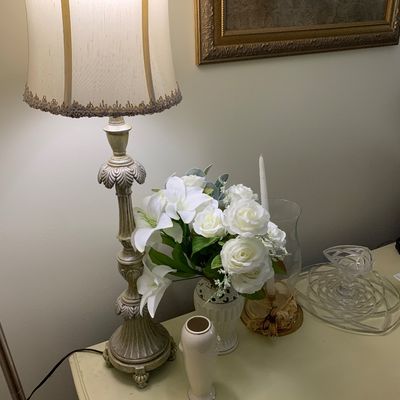 Sale Photo Thumbnail #10: Traditional table lamp with an ornate, distressed-finish base and a fringed fabric shade, shown alongside a faux floral arrangement in a white vase and other tabletop decor.
