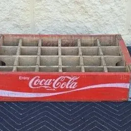 Vintage red wooden Coca-Cola bottle crate with metal-reinforced corners and 24-bottle dividers, showing some wear and fading from age.