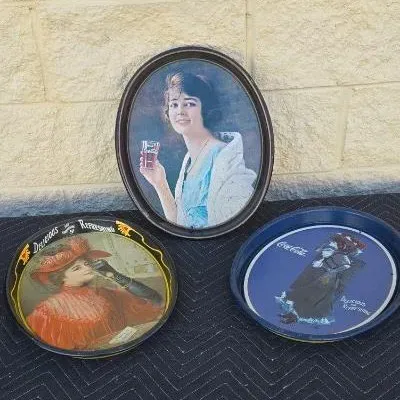 Three vintage Coca-Cola metal serving trays, including one oval tray featuring a woman in a blue dress and two round trays with classic...
