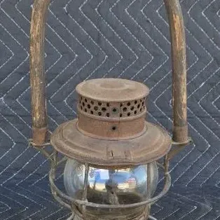 Vintage railroad lantern featuring a rusted metal frame, clear glass globe, and a tall, curved wooden carrying handle.