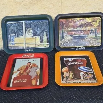 Four vintage Coca-Cola metal serving trays featuring various retro advertisements and illustrations, showing some light wear and scratches.