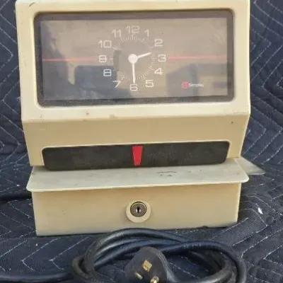 Vintage Simplex industrial time clock with a beige plastic casing, analog clock face, and attached power cord.