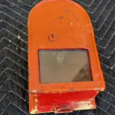 Vintage orange painted metal coin bank with a glass viewing window and keyhole, showing some wear and paint loss.