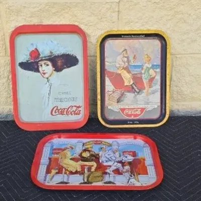 Three vintage Coca-Cola metal serving trays featuring classic advertising illustrations, showing some wear and patina consistent with age.