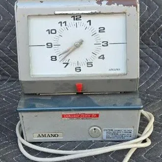 Vintage Amano industrial time clock with a grey metal housing, showing some wear and paint loss, complete with its power cord.