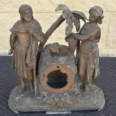 Antique cast metal clock case featuring a man and woman by a well under a palm tree, showing significant wear and missing the clock...