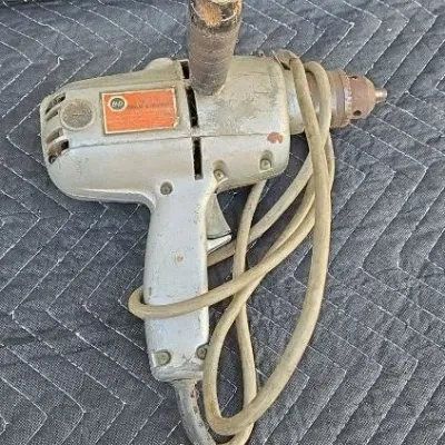 Vintage Black & Decker heavy-duty electric drill with a silver metal casing, top auxiliary handle, and attached power cord.