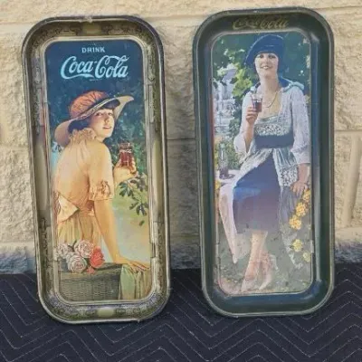 Pair of vintage Coca-Cola metal serving trays featuring classic illustrations of women in early 20th-century attire, showing some wear...