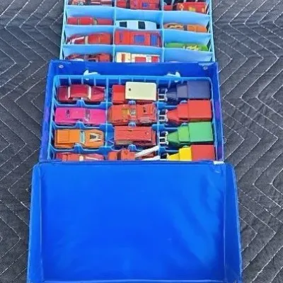 Vintage blue vinyl Matchbox or Hot Wheels carrying case filled with various die-cast toy cars and trucks in plastic organizer trays.
