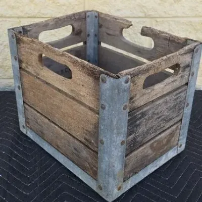 Vintage wooden milk crate with galvanized metal corner brackets and integrated handle cutouts, showing rustic wear and weathering.