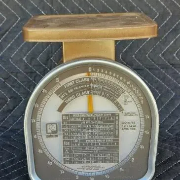 Vintage Pelouze Model Y5 postal scale with a gold-toned metal body and detailed rate chart on the face, dated April 1988.