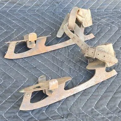 Pair of antique clamp-on ice skates with rusted metal blades and worn leather heel straps, showing significant age and patina.
